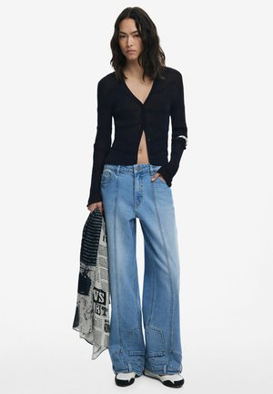 Schwarze gerippte Langarm-Cardigan mit V-Ausschnitt und Knöpfen, kombiniert mit hellblauen, weit geschnittenen Jeans. Hält einen gemusterten Schal in einer Hand.
