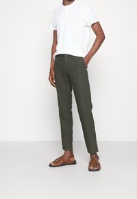 Pantaloni verde oliva in misto lino dal taglio slim, abbinati a una maglietta henley bianca. Sandali con cinturino marroni completano l'outfit.
