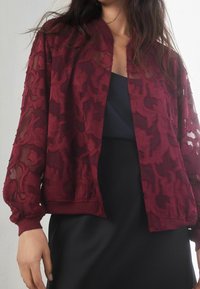 Blouson aviateur transparent bordeaux avec texture en dentelle florale, poignets et ourlet côtelés. Porté sur un haut foncé, avec une coupe décontractée.