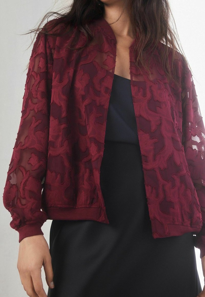 Blouson aviateur transparent bordeaux avec texture en dentelle florale, poignets et ourlet côtelés. Porté sur un haut foncé, avec une coupe décontractée.