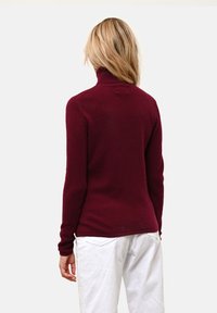 Burgundy polotröja i mjukt material. Har långa ärmar och en figurnära design, visad bakifrån mot en enkel bakgrund.