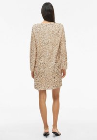 Sukienka z długim rękawem pokryta jasnobeżowymi cekinami, o długości do kolan, prostym kroju, z okrągłym dekoltem, o fakturowanej powierzchni, noszona z sandałami na odkryte palce.