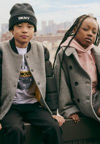 Dos niños vestidos con ropa exterior gris y ropa de marca DKNY, sentados al aire libre con un horizonte urbano de fondo.