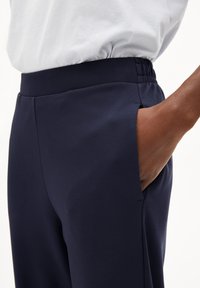 Pantaloni blu navy con una texture liscia, dotati di una vita elastica e tasche laterali. Dettaglio ravvicinato della tasca e della caduta del tessuto.