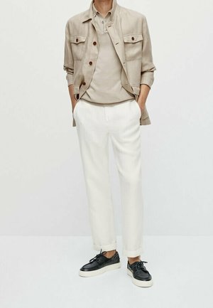Light beige linen jacket with two front pockets over a matching beige shirt, paired with white tapered trousers and black sneakers.