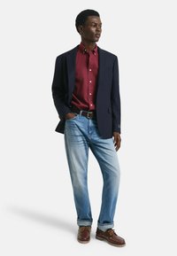Navyblazer über einem burgunderfarbenen Hemd mit Knöpfen, kombiniert mit hellblauen Jeans und braunen Schuhen. Einfaches Design mit klassischer Passform und subtilen Texturen.