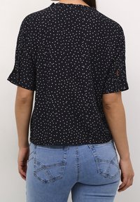 Top nero a maniche corte con pois bianchi, caratterizzato da un colletto arricciato e dettaglio di bottoni sulle maniche. Indossato con jeans blu, vista posteriore.