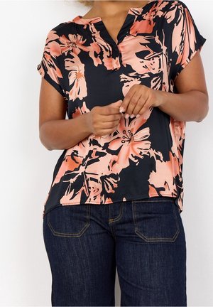 Donna che indossa una maglietta nera a maniche corte con grande stampa floreale pesca e jeans blu scuro, mani intrecciate in vita.