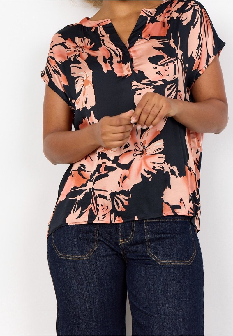 Femme portant un haut noir à manches courtes avec un grand imprimé floral pêche et un jean bleu foncé, les mains jointes à la taille.