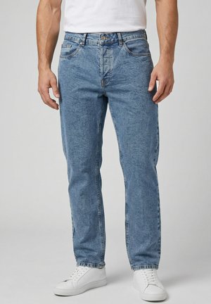 Mand iført lyseblå jeans med lige ben, hvide sneakers og en hvid tætsiddende t-shirt, stående mod en ensfarvet baggrund.