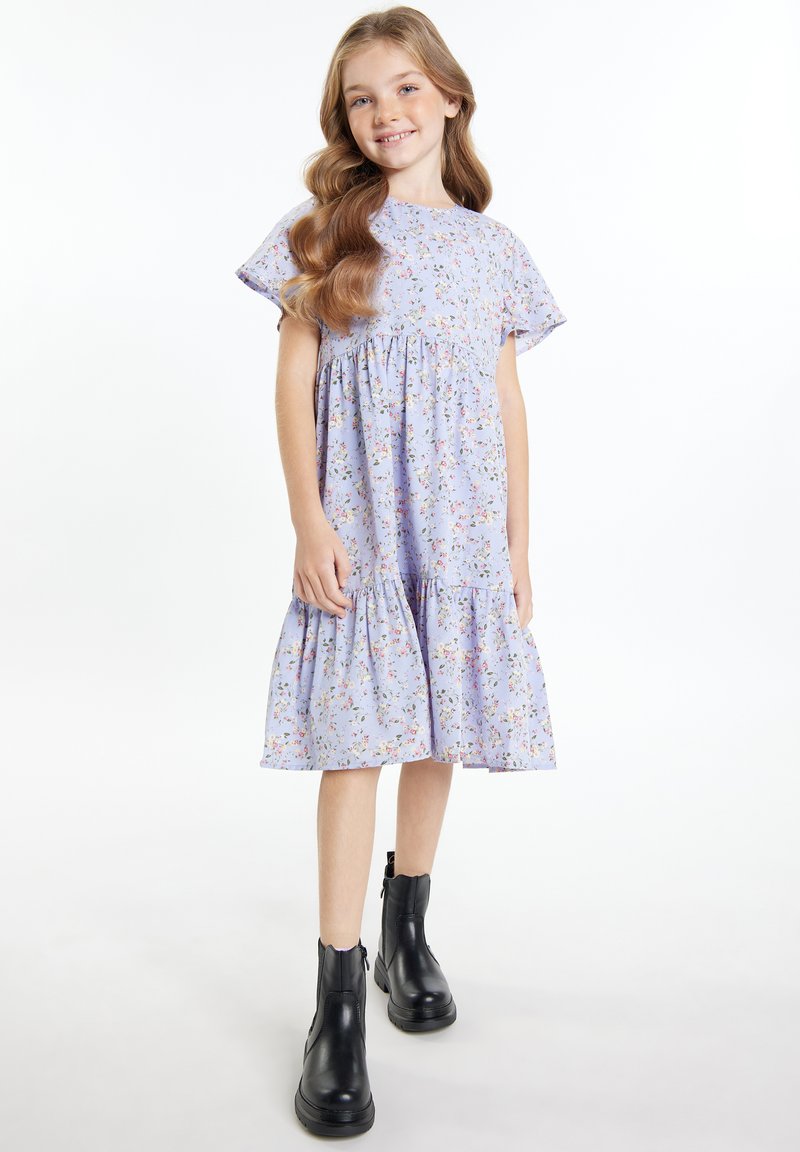 Lila Blumenkleid mit kurzen, ausgestellten Ärmeln und einem gestuften Saum, kombiniert mit schwarzen Stiefeletten. Weicher Stoff mit leichter Textur.