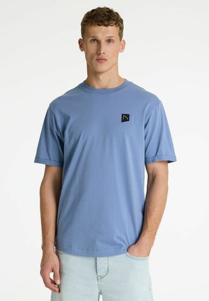 Mężczyzna ubrany w jasnoniebieską koszulkę z krótkim rękawem z małą czarną naszywką z logo na piersi oraz jasne dżinsy.