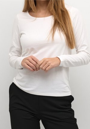 Femme aux longs cheveux châtain clair portant un haut blanc uni à manches longues et un pantalon noir, mains jointes légèrement devant.