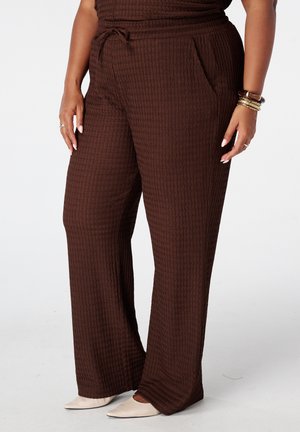 Femme portant un pantalon large marron texturé avec des poches, associé à des chaussures pointues crème et des bracelets dorés au poignet.