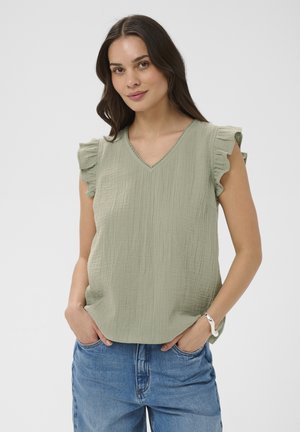 Woman with brown hair wearing light green ruffled sleeve blouse and blue jeans, hands in pockets, smiling gently against plain background.