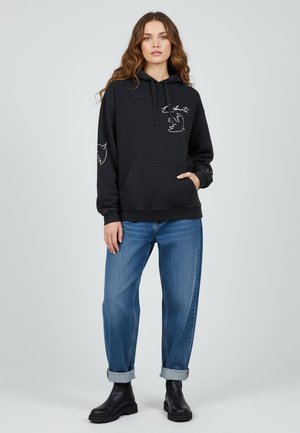 Haut à capuche noir avec poche avant, cordon de serrage à la capuche et détails graphiques blancs. Associé à un jean bleu retroussé et des bottines noires.