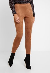 Femme portant un pantalon en suède marron taille haute, associé à des bottines chaussettes à talons hauts noires à bouts pointus et un haut à manches longues imprimé léopard.