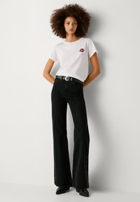 Fehér póló piros ajak grafikával, magas derekú fekete harangnadrággal, szemlyuk részletekkel és tüskés fekete övvel párosítva.