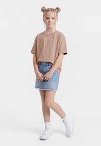 Mlada deklica z blond kodrastimi opravljenimi figami stoji oblečena v bež majico, jeans krilo, bele nogavice in bele športne superge na beli podlagi.