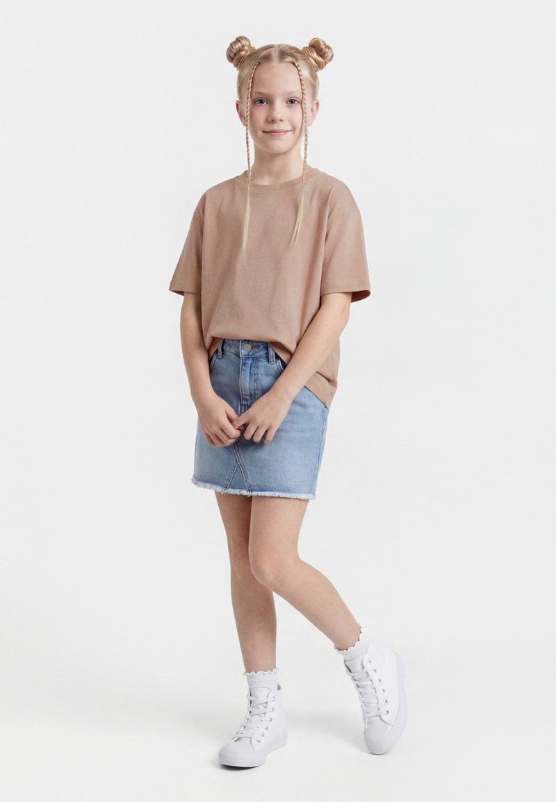 Mlada deklica z blond kodrastimi opravljenimi figami stoji oblečena v bež majico, jeans krilo, bele nogavice in bele športne superge na beli podlagi.