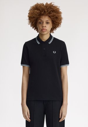 Kiharatukkainen nainen, joka on pukeutunut mustaan lyhythihaiseen poolopaitaan, jossa on valkoiset raidalliset kaulus ja hihansuut, seisoo yksinkertaisen valkoisen taustan edessä.