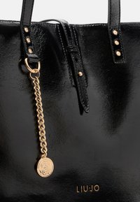 Borsa tote in pelle ecologica nera con finitura lucida, dotata di hardware color oro, dettaglio a catena, piccolo medaglione e tre borchie decorative sulle cinghie.