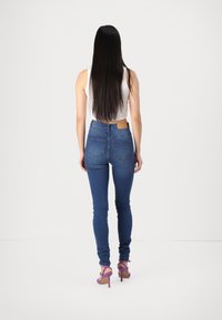 Femme aux longs cheveux noirs portant un haut blanc sans manches, un jean skinny bleu et des sandales à talons hauts violettes, debout de dos.