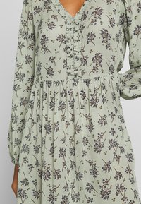 Robe vert clair à manches longues avec motif floral violet foncé et marron et décolleté en V froufroutant, portée par une personne avec une main sur la hanche.