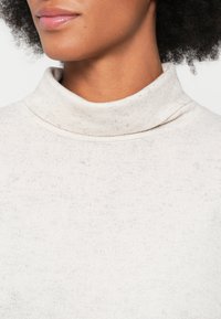 Close-up of a person’s neck and lower face wearing a light beige turtleneck sweater with natural textured hair.