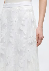 Λευκή φούστα με μεγάλα κεντημένα λουλουδένια σχέδια και διακριτικά παγιέτες, φορεμένη από ένα άτομο με γυμνό μπράτσο που φαίνεται μπροστά σε απλό φόντο.
