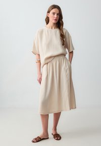 Haut beige léger à manches courtes avec un col rond, associé à une jupe midi fluide à taille élastique assortie. Sandales brunes à enfiler.