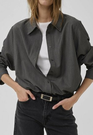 Persona que lleva una camisa oscura de rayas finas abotonada sobre una camiseta blanca, con vaqueros negros y un cinturón negro, con las manos metidas en los bolsillos delanteros.