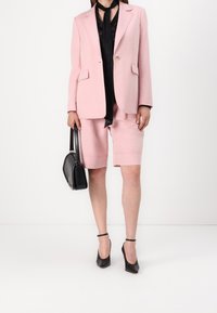 Blazer rose pâle sur mesure avec deux poches frontales, associé à un short assorti. Écharpe en soie noire et sac à main noir. Talons noirs complètent le look.