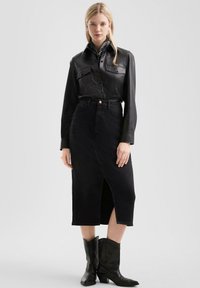 Zwart leren shirt met twee borstzakken, gecombineerd met een zwarte spijkerrok met hoge taille en een split aan de voorkant. Zwarte cowboylaarzen maken de look compleet.