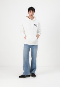 Sweat-shirt à capuche blanc avec poche avant et logo, associé à un jean évasé bleu clair. Modèle portant des chaussures noires, se tenant contre un fond blanc.