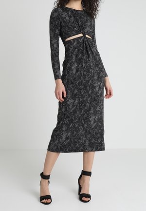 Kobieta w czarno-szarej marmurkowej sukience midi z długimi rękawami i wycięciami na talii, w połączeniu z czarnymi sandałami na wysokim obcasie.