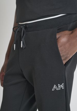 Close-up van een persoon die zwarte joggers draagt met een trekkoord in de taille en een wit "AM" logo bij de rechterzak, hand gedeeltelijk in de zak.