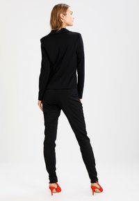 Svart skräddarsydd kavaj med en figursydd design, kombinerad med slim fit svarta byxor, bärs med röda högklackade skor. Slät tygstruktur.