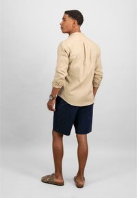 Camicia a maniche lunghe beige chiaro con maniche a tab, abbinata a pantaloni corti blu navy e sandali marroni, vista da dietro. Design semplice e vestibilità comoda.
