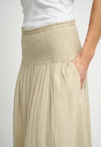 Gonna maxi di lino beige con vita arricciata, tasche laterali e pieghe verticali per texture e movimento.