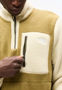 Άτομο που φοράει μπουφάν από φλις σε μπεζ και εκρού χρώμα με φερμουάρ μπροστά και τσέπη στο στήθος με φερμουάρ, τραβώντας το φερμουάρ προς τα κάτω.