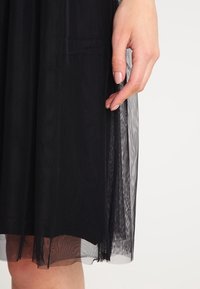 Svart tyllkjol med ett lagerdesign, lätt textur, och en hand som syns hålla i kanten. Slutar precis ovanför knäet.