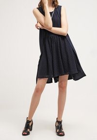 Robe sans manches bleu marine en tissu texturé, jupe évasée et dos plus long. Associée à des sandales à talons en cuir noir avec des lanières.