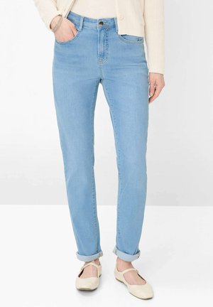 Frau trägt hellblaue hochgekrempelte Jeans, cremefarbigen Cardigan, beigefarbene Mary Jane Ballerinas, mit einer Hand in der Tasche und Armbändern am Handgelenk.