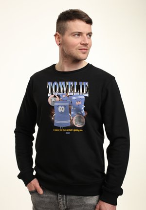 Mężczyzna z krótkimi włosami noszący czarną bluzę z animowanymi niebieskimi ręcznikami i napisami „Towelie” oraz „Nie mam pojęcia, co się dzieje.”