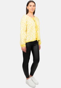 Geel vest met wit luipaardprint, knoopsluiting en strikdetail. Gekozen in combinatie met zwarte leggings en witte sneakers, wat een casual uitstraling geeft.