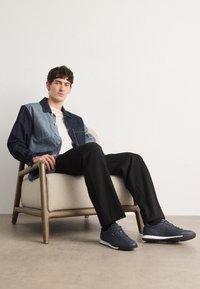 Veste en denim avec des panneaux bleu clair et bleu foncé, portée sur une chemise blanche. Pantalon noir et baskets foncées complètent la tenue. Assis sur une chaise en bois.