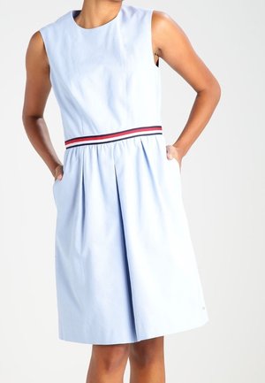 Světle modré šaty bez rukávů s kulatým výstřihem, pas s červenými a tmavě modrými pruhy a plisovanou sukní s bočními kapsami.