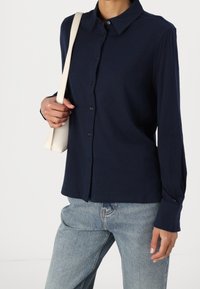 Granatowa koszula zapinana na guziki, wykonana z miękkiego materiału, z klasycznym kołnierzykiem i długimi rękawami. Połączona z jasnoniebieskimi dżinsami i kremową torbą.