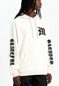 Felpa crema, materiale in cotone morbido, ampio tascone frontale, testo nero stampato sulle maniche ("M," "GRATEFUL," "BLESSED"). Polsini a coste.